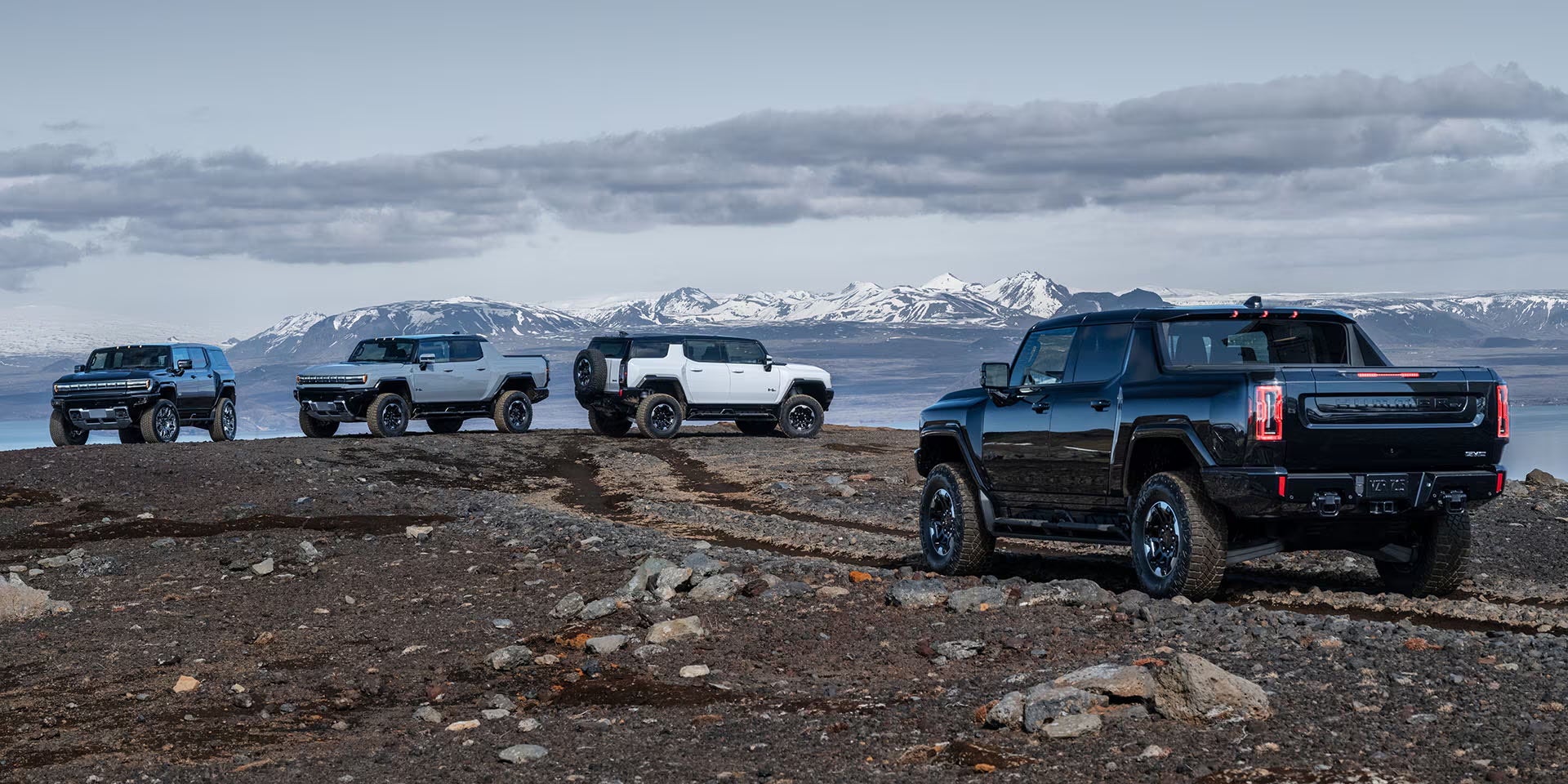 Four GMC Hummer EVs parked on a rocky hilltop with snow-covered mountains in the background.