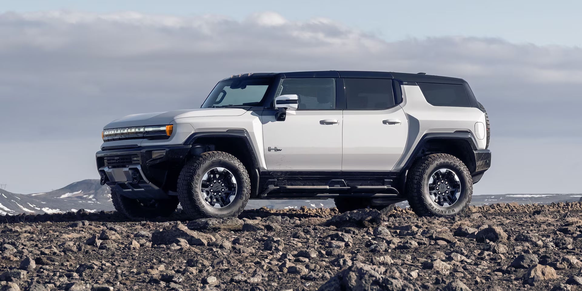 White GMC Hummer EV SUV parked on a rocky landscape with snowy mountains in the distance.