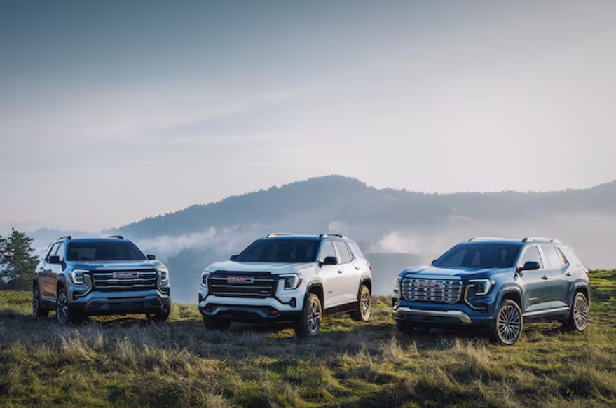 Three 2026 GMC Terrain SUVs parked on a grassy hilltop with mountains and fog in the background.