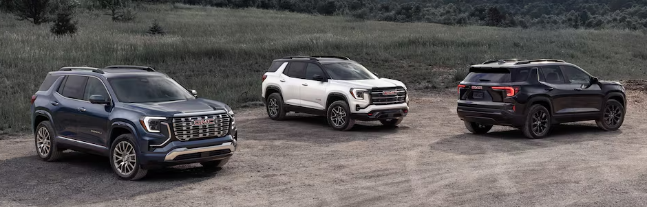 Three 2026 GMC Terrain SUVs in blue, white, and black parked on a dirt lot near grassy fields.